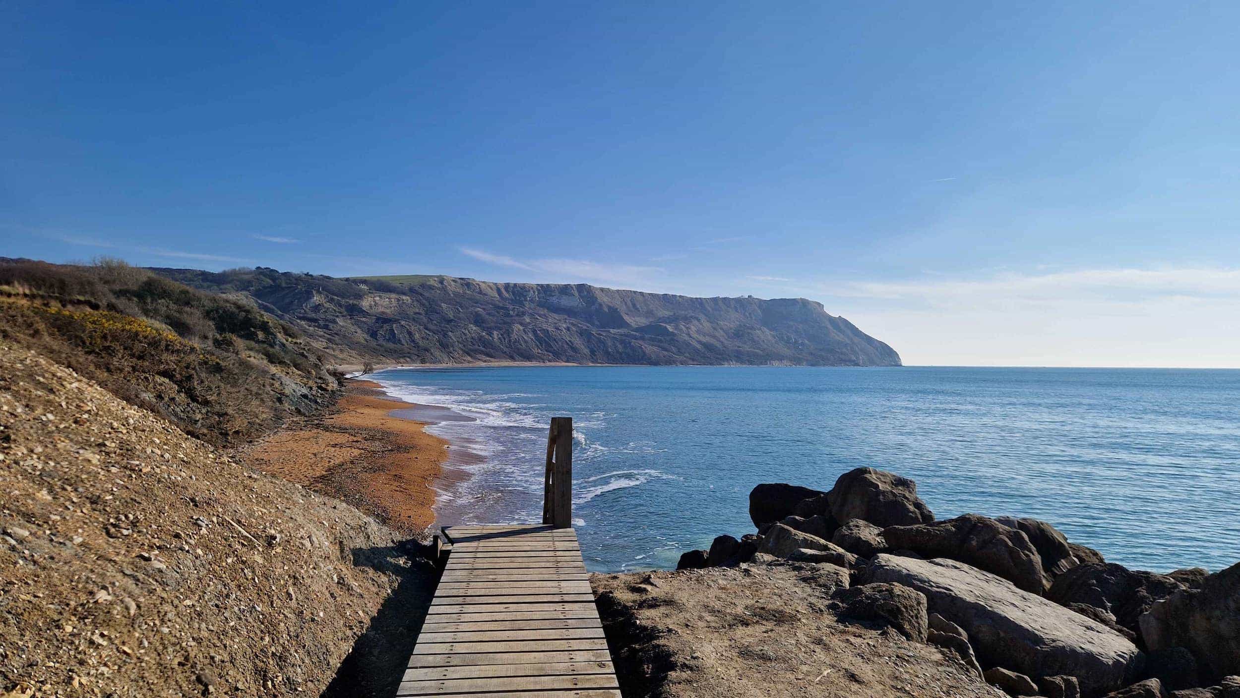 Wooden boardwalk leading to a rocky coastline with clear blue water, backed by steep cliffs and a sandy beach under a bright blue sky.